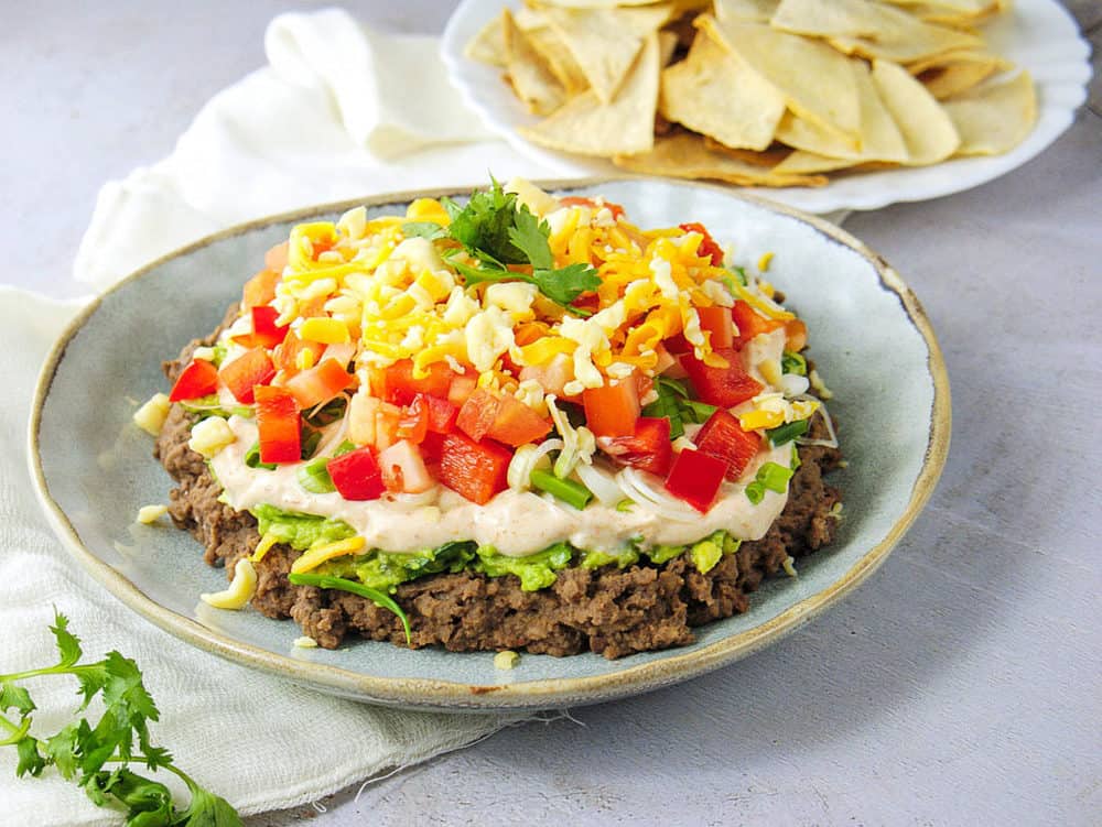 healthy 7 layer taco dip made with beans, avocado, greek yogurt, veggies and cheese, pictured on a blue plate with chips in the background.