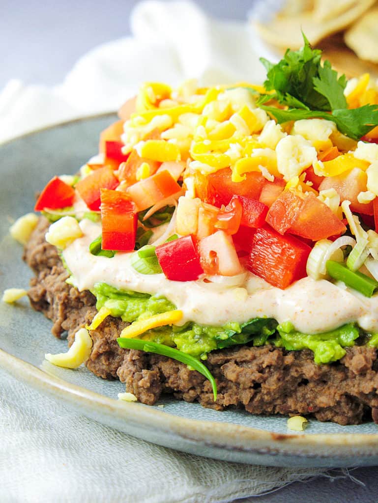 closeup of healthy 7 layer taco dip made with beans, avocado, greek yogurt, veggies and cheese, pictured on a blue plate.