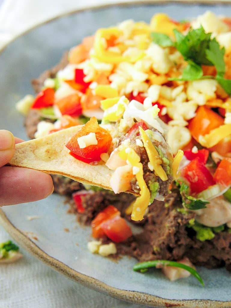 closeup of healthy 7 layer taco dip made with beans, avocado, greek yogurt, veggies and cheese, pictured on a blue plate, with a chip dipped in it.