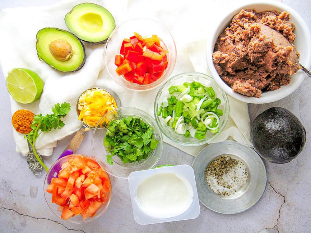 ingredients for healthy 7 layer taco dip: refried beans, tomatoes, bell peppers, green onions, cilantro, avocado, cheese, pictured on a marble background, top view