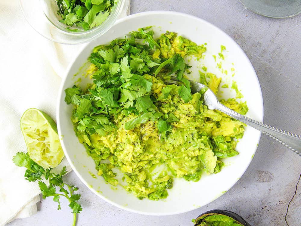 avocado mashed with lime juice, cilantro, salt, pepper, in a white bowl