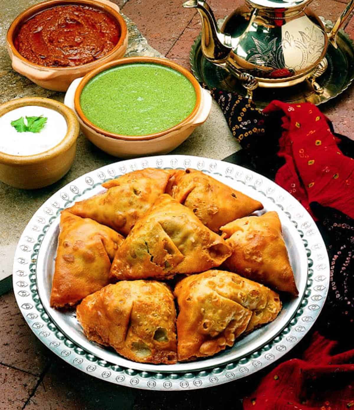 Indian samosas on a white plate with green and red chutney in the background.