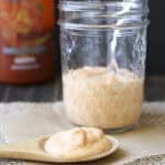 Wooden spoon filled with creamy sriracha sauce laying on parchment in front of glass sauce jar