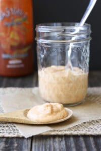 Wooden spoon filled with creamy sriracha sauce laying on parchment in front of glass sauce jar