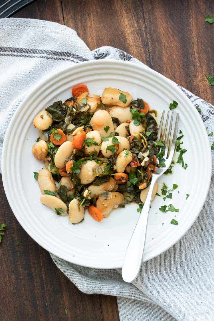 This healthy lima bean recipe with added veggies is a one stop meal. Gigantes Plaki is loaded with protein, perfect for all ages and so flavorful! #limabeanrecipes #greekrecipes Baked lima beans and veggies on a white plate