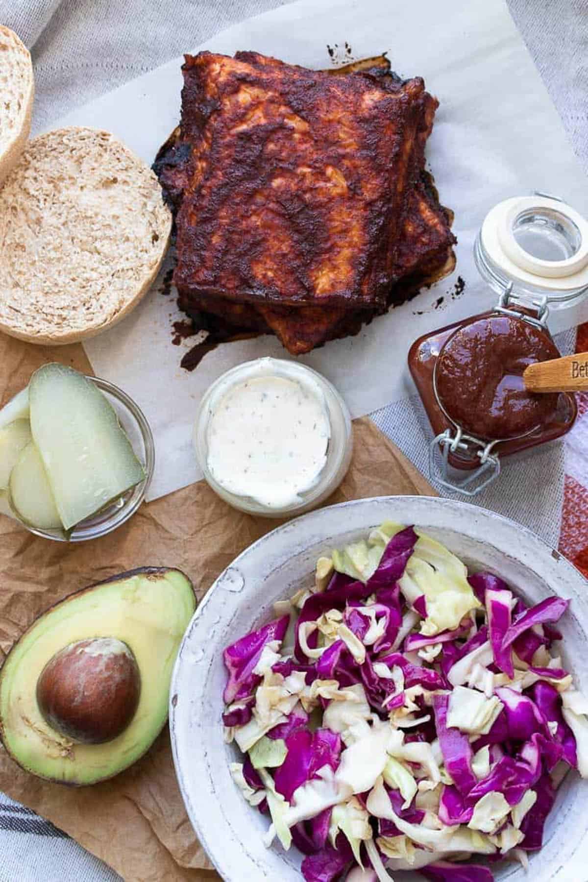 Bread roll, BBQ tempeh squares, shredded cabbage, avocado, pickles, ranch and BBQ sauce on a table.