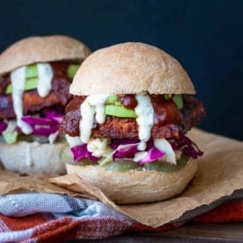 Front view of a BBQ tempeh sandwich in a bread roll with cabbage, avocado, pickles and ranch, on a cutting board.