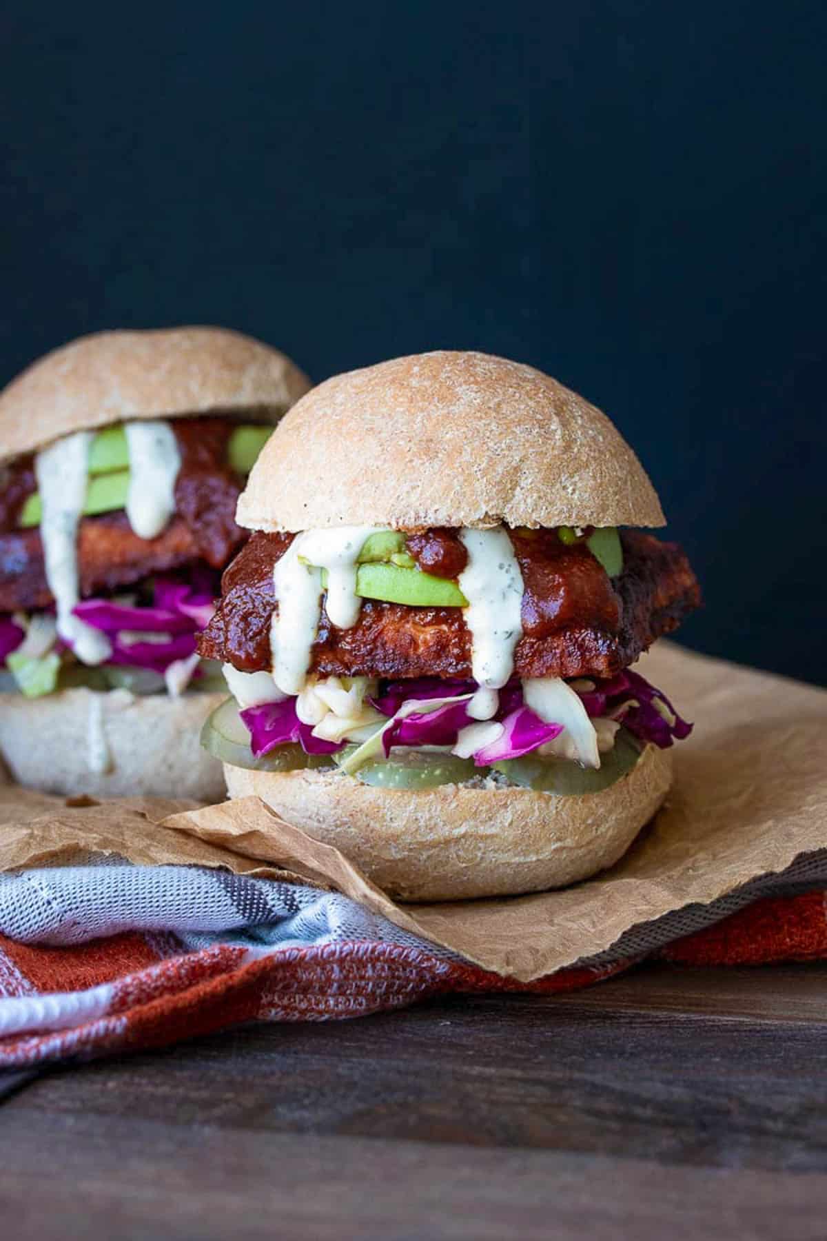 Front view of a BBQ tempeh sandwich in a bread roll with cabbage, avocado, pickles and ranch, on a cutting board.