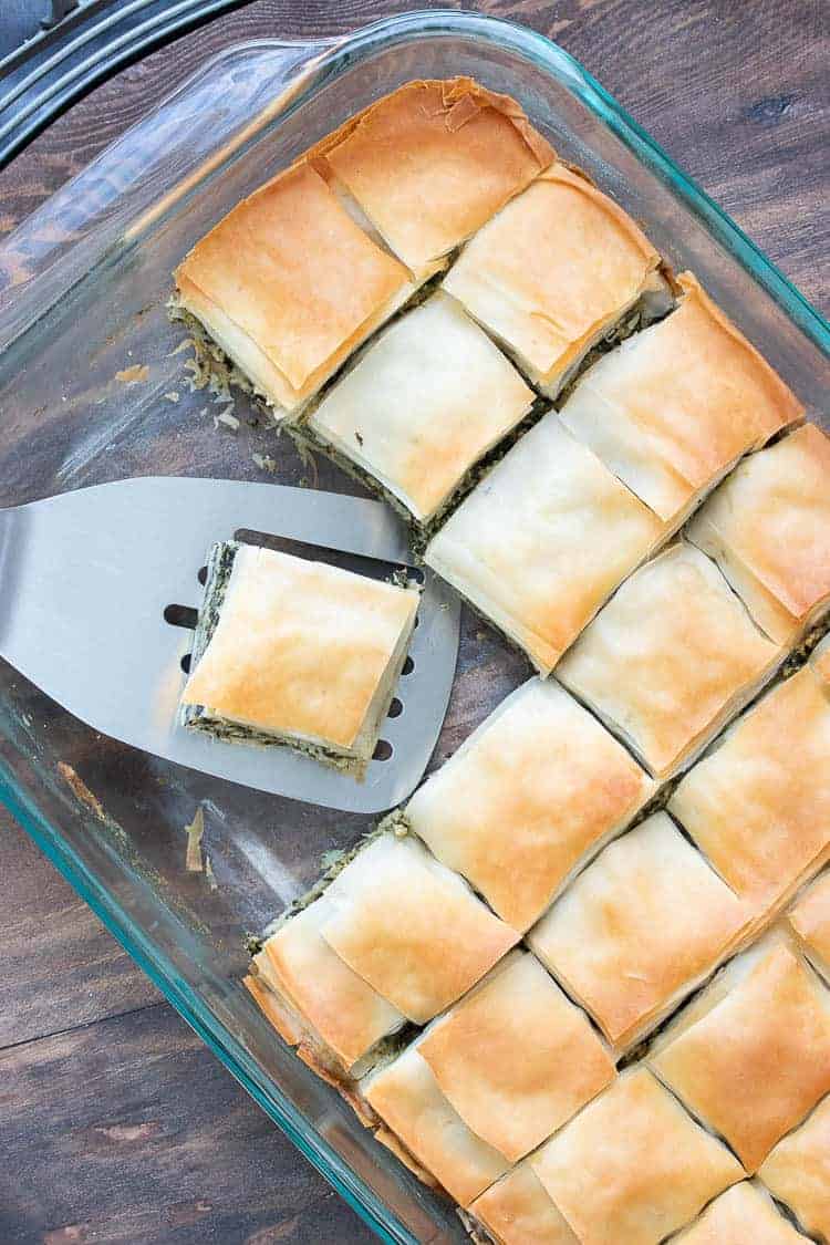 This authentic Greek recipe has gotten a makeover and is better than ever. This vegan spanakopita is hands down the absolute best you've ever had! #veganappetizers #greekrecipes Spatula picking up a piece of baked spanakopita from a glass baking dish