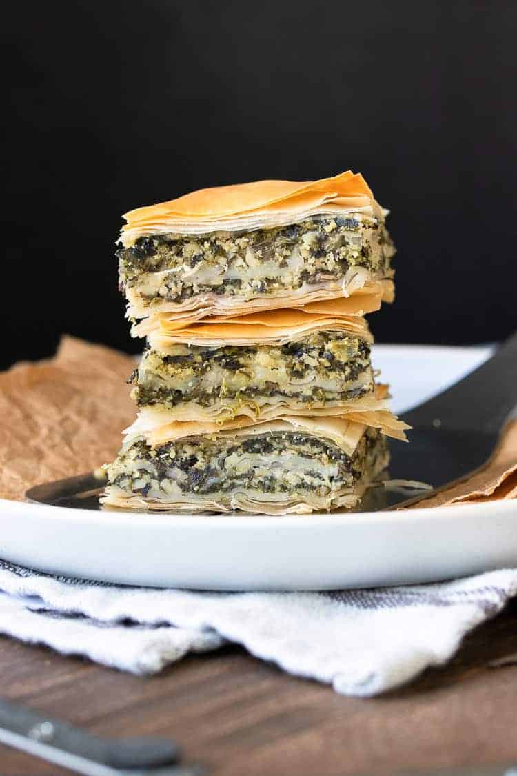 This authentic Greek recipe has gotten a makeover and is better than ever. This vegan spanakopita is hands down the absolute best you've ever had! #veganappetizers #greekrecipes Pile of three pieces of spanakopita on a white plate