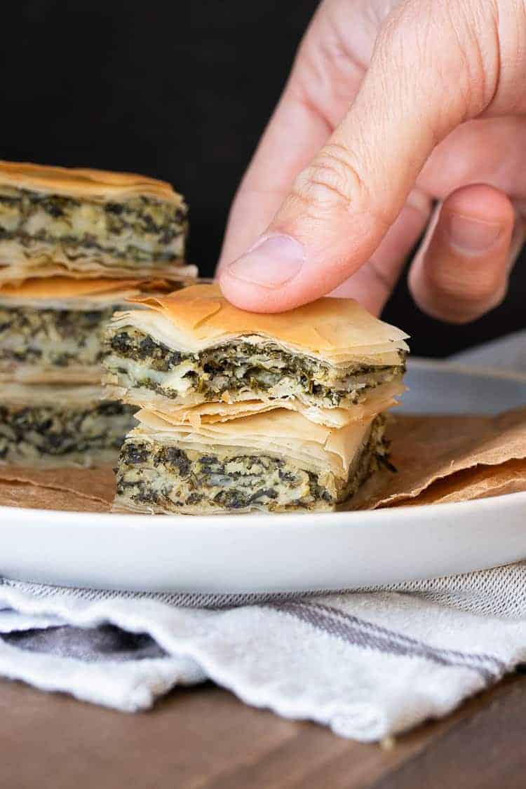 This authentic Greek recipe has gotten a makeover and is better than ever. This vegan spanakopita is hands down the absolute best you've ever had! #veganappetizers #greekrecipes Hand picking up a piece of spanakopita with a bite out of it