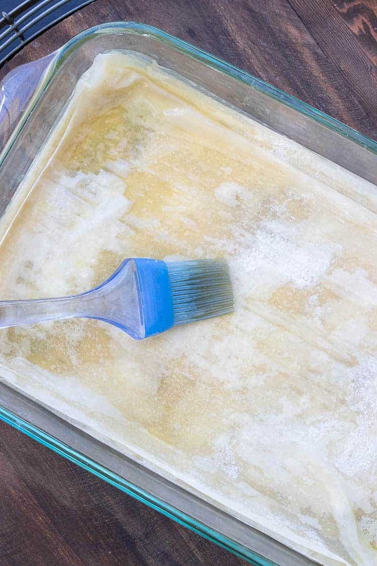 This authentic Greek recipe has gotten a makeover and is better than ever. This vegan spanakopita is hands down the absolute best you've ever had! #veganappetizers #greekrecipes Blue pastry brush brushing olive oil on a phyllo (filo) pastry sheet