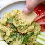 Hand dipping pita chip into a pile of hummus topped with oil and parsley.