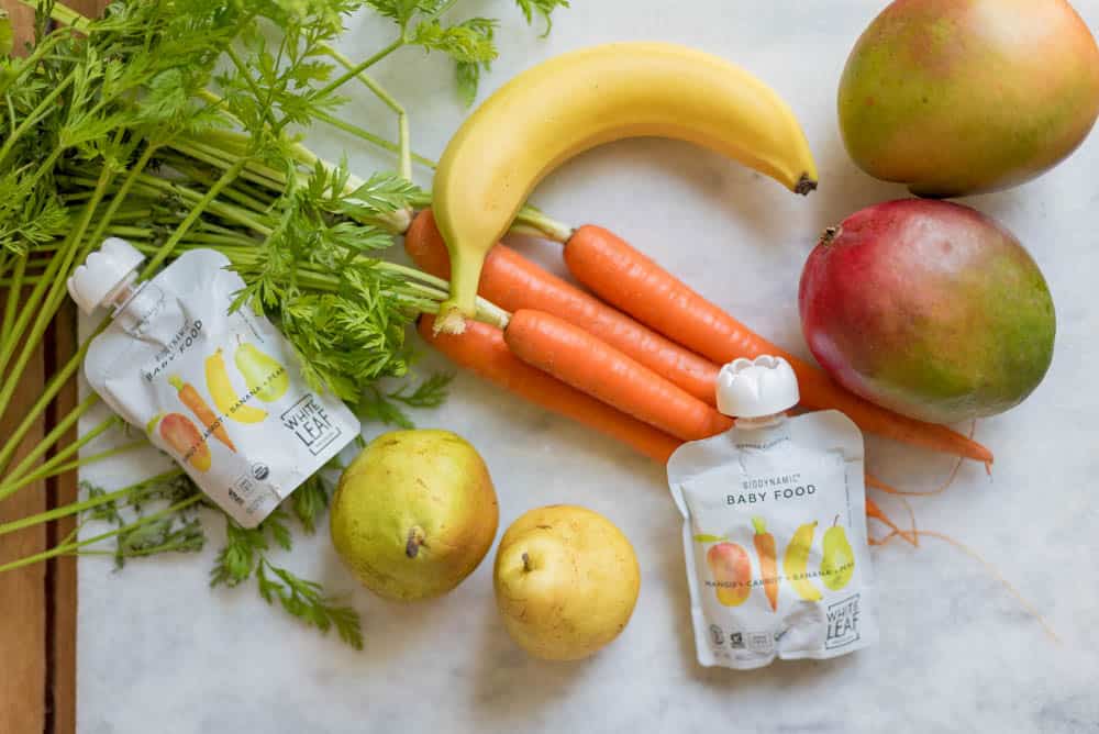 white leaf provisions baby food pouches on a white backdrop, surrounded by fresh mangoes, bananas, pears, carrots