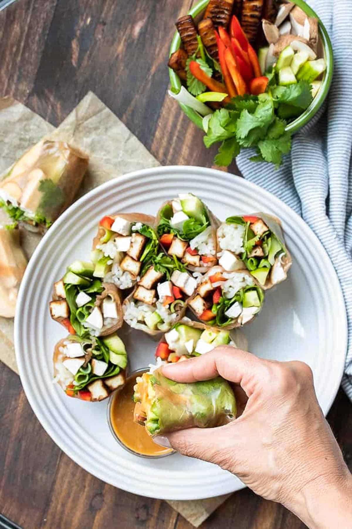 Hand dipping half a vegetable summer roll into brown sauce on a white plate.