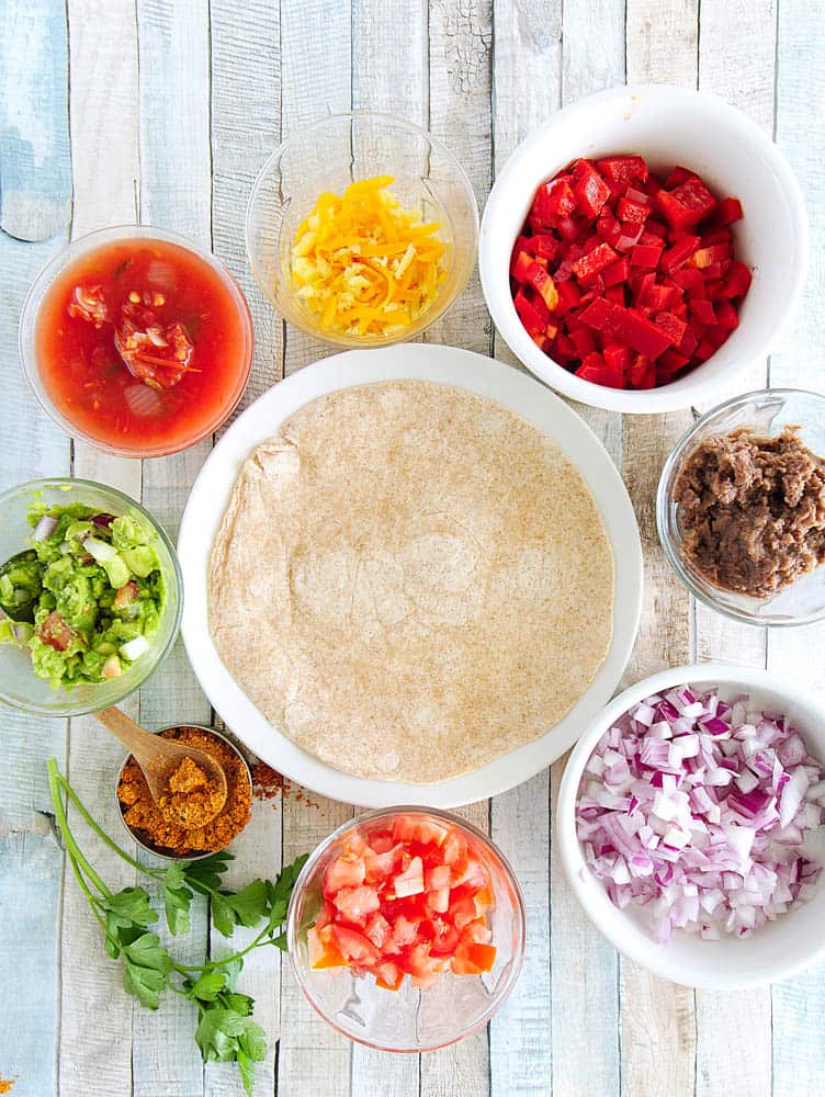 ingredients for mexican pizza: tortilla, cheese, bell pepper, taco seasoning, salsa, guacamole, tomatoes, onions, refried black beans