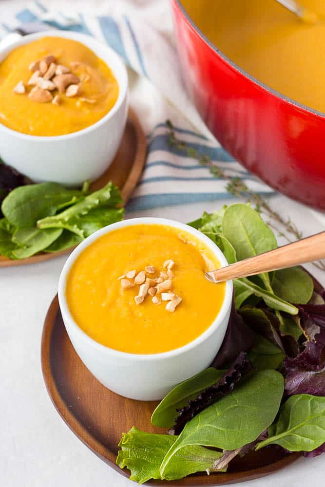 Butternut squash soup in small white bowl topped with nuts and a on a plate with a side of greens.