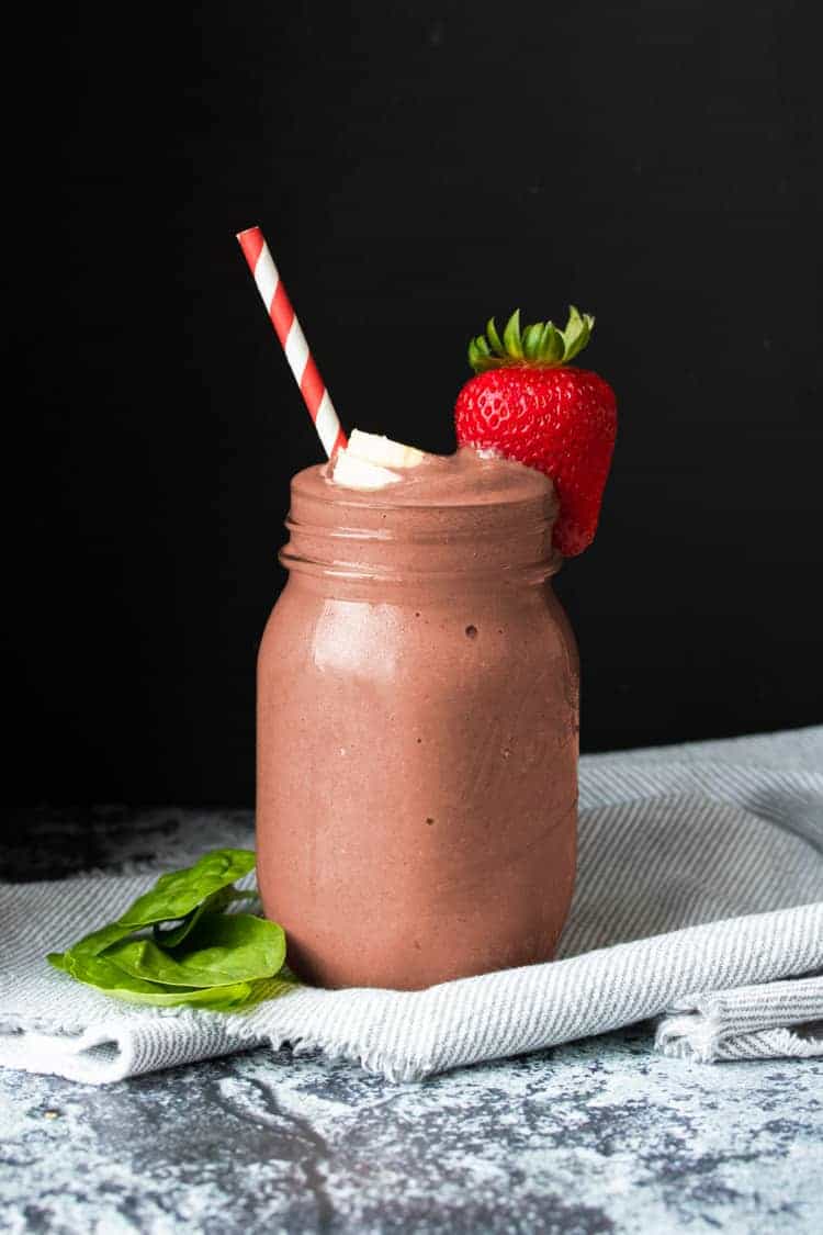 Start your day with this healthy strawberry spinach smoothie with chia seeds for added thickness. The added low fat vegan protein helps keep you full and energized all morning long! #healthysmoothies #veganbreakfast #ad Glass jar with a reddish smoothie surrounded by pieces of strawberry, banana and spinach
