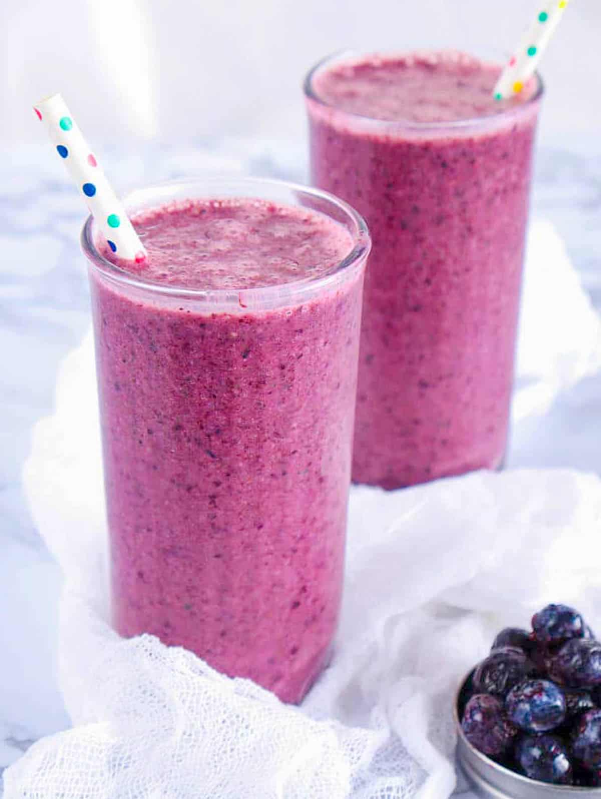 Blueberry pineapple smoothie in two glasses with a straw against a grey background.
