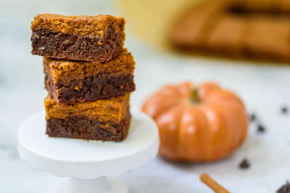 layered pumpkin brownies stacked on a white pedestal, with cinnamon, chocolate chips and pumpkin in the background