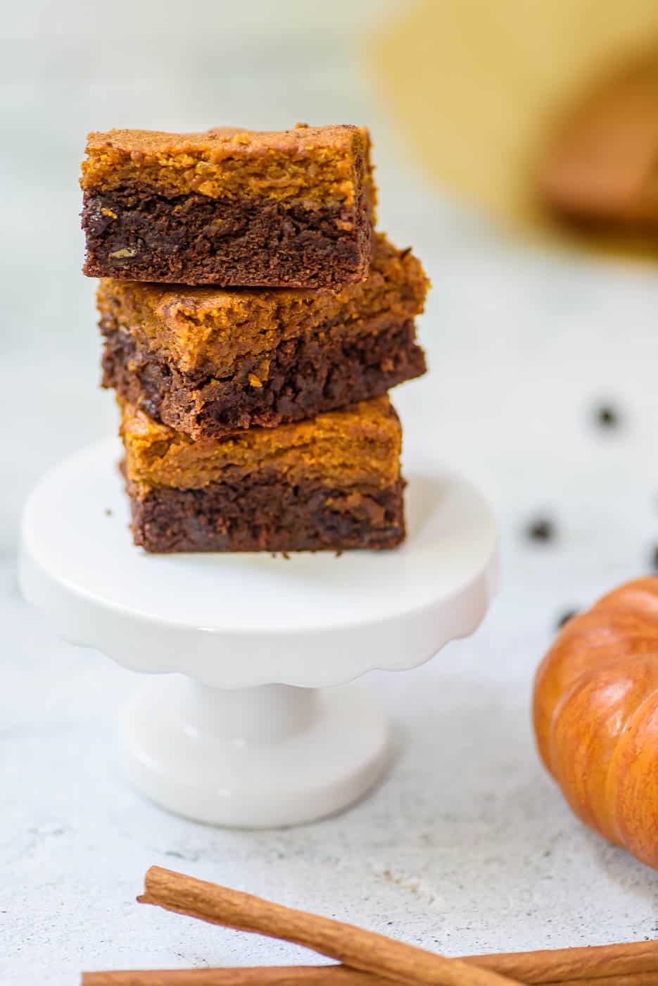 healthy pumpkin brownies stacked on a white pedestal, with cinnamon, chocolate chips and pumpkin in the background 