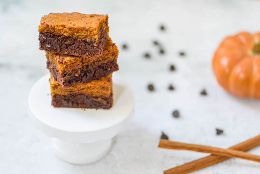 layered pumpkin brownies stacked on a white pedestal, with cinnamon, chocolate chips and pumpkin in the background
