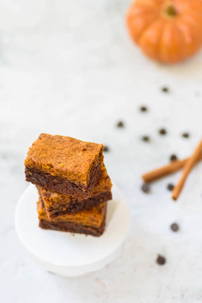 layered pumpkin brownies stacked on a white pedestal, with cinnamon, chocolate chips and pumpkin in the background