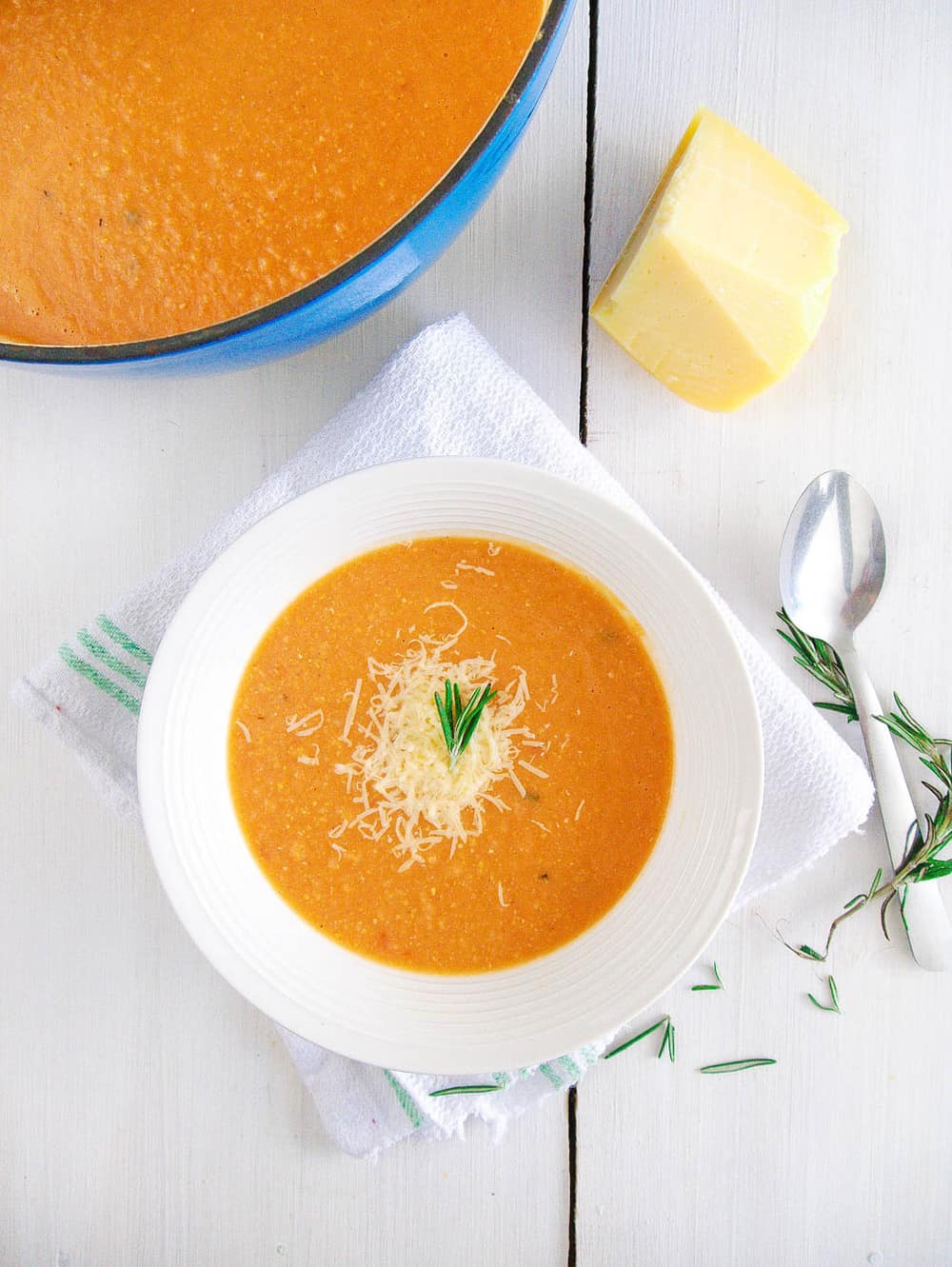 A white bowl of garbanzo bean soup with parmesan and rosemary on a white background with rosemary and a spoon.