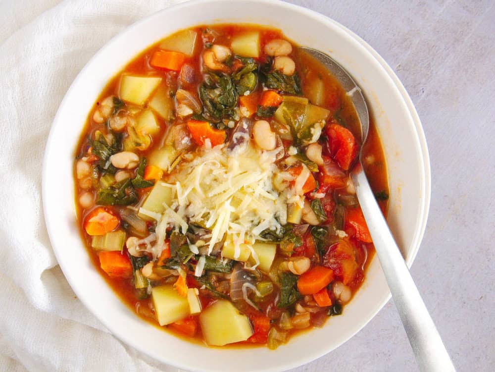 healthy minestrone soup in a white bowl with a spoon, topped with shredded parmesan cheese