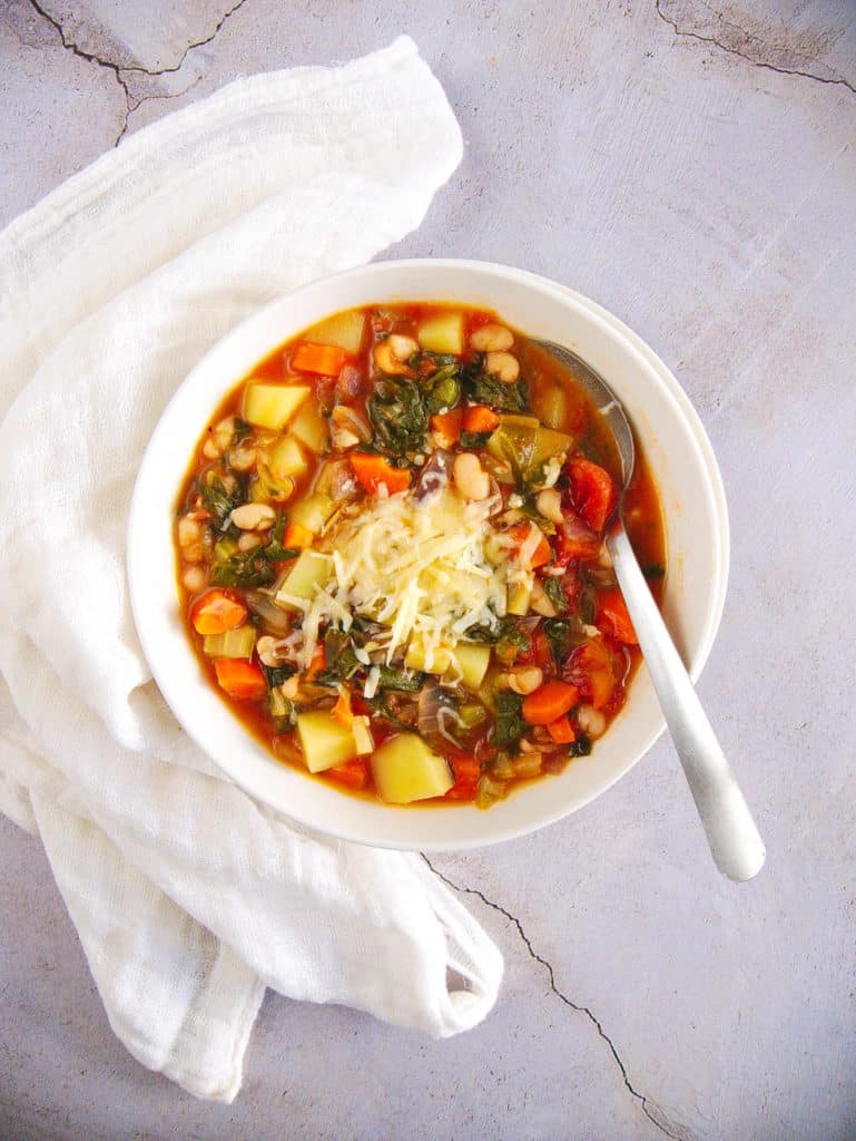 healthy minestrone soup in a white bowl with a spoon, topped with shredded parmesan cheese