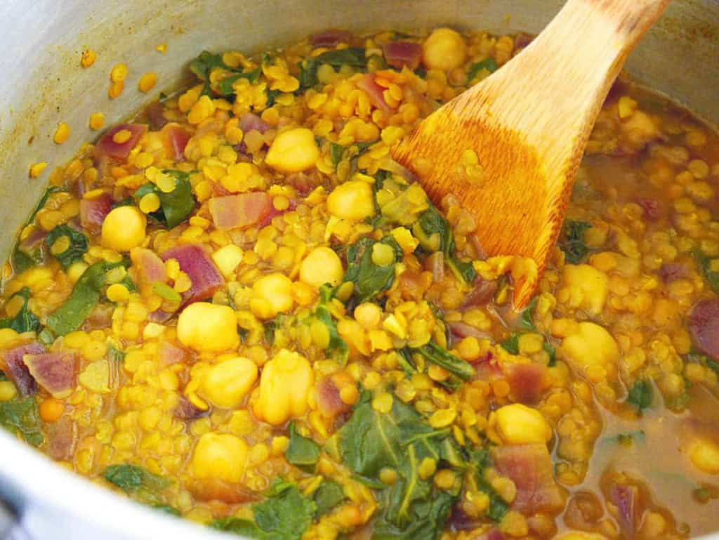 Wooden spoon stirring chickpeas lentils and swiss chard in a large pot with spices.