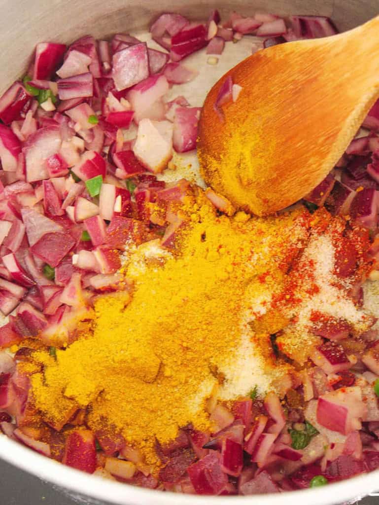 Spices added to a pot with onions and other herbs, mixed with a wooden spoon.