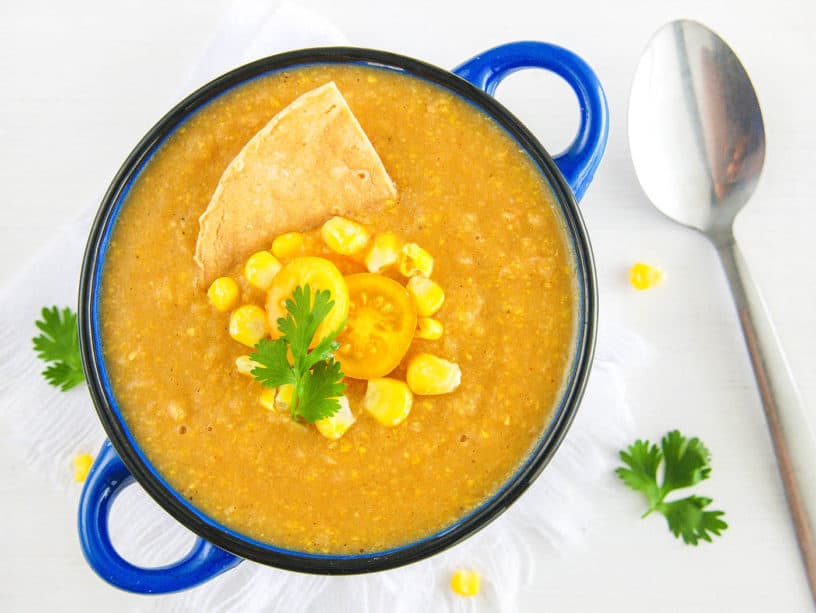 potato corn chowder - spicy, dairy-free, served in a blue bowl with fresh corn, tomatoes, and a corn chip as garnishes