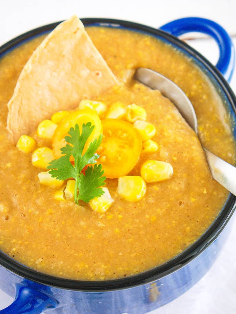potato corn chowder - spicy, dairy-free, served in a blue bowl with fresh corn, tomatoes, and a corn chip as garnishes with a spoon in the bowl