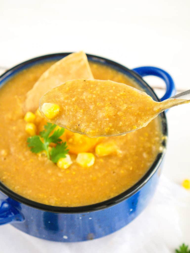 a spoonful of potato corn chowder - spicy, dairy-free, served in a blue bowl with fresh corn, tomatoes, and a corn chip as garnishes