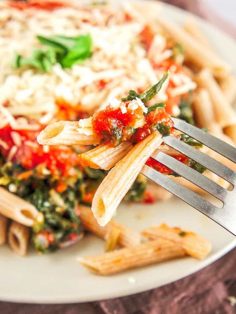 bite of whole wheat pasta recipe topped with spinach, pasta sauce, parmesan cheese on a white plate