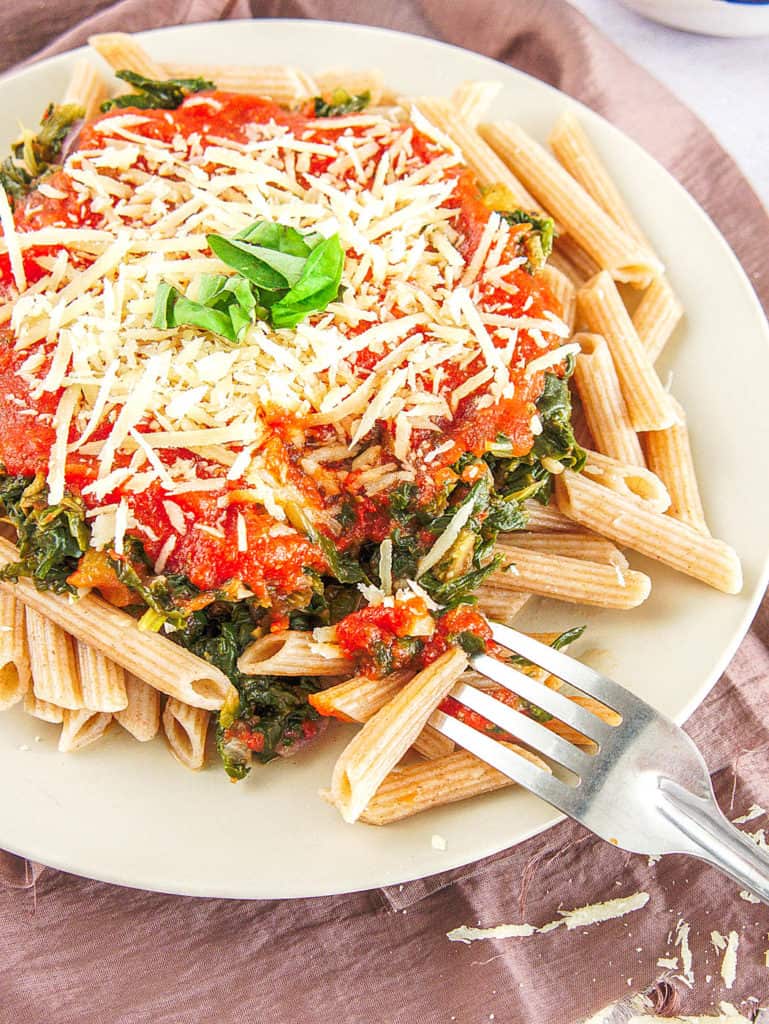whole wheat pasta recipe topped with spinach, pasta sauce, parmesan cheese on a white plate, with a fork