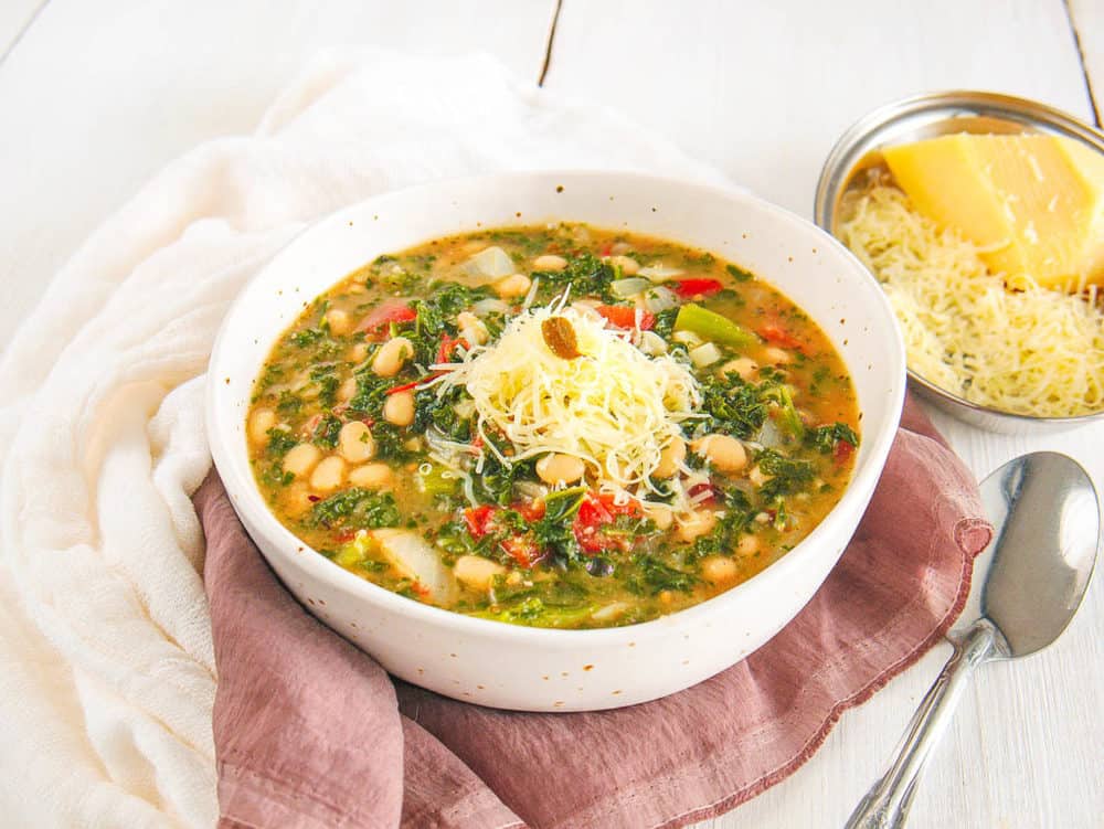 white bean and kale soup, served in a white bowl, topped with parmesan cheese with spoon on the side