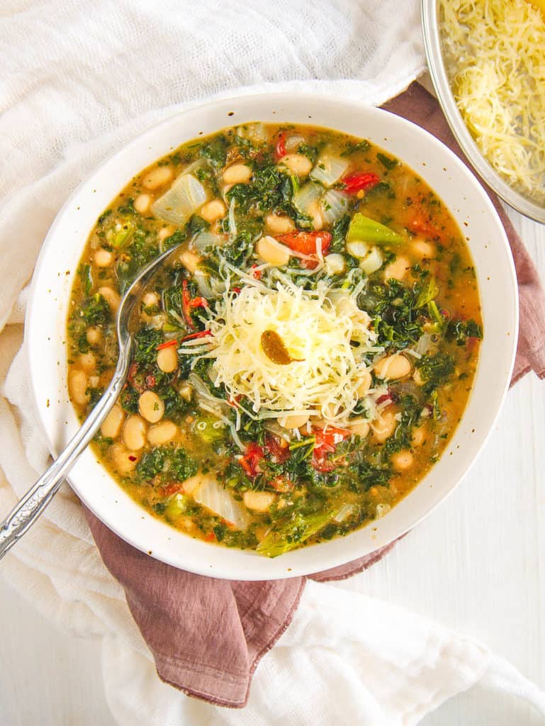 white bean and kale soup, served in a white bowl, topped with parmesan cheese and spoon - top vie