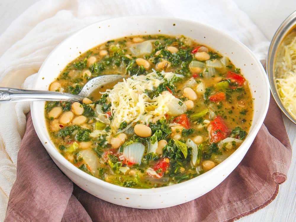 white bean and kale soup, served in a white bowl, topped with parmesan cheese with spoon