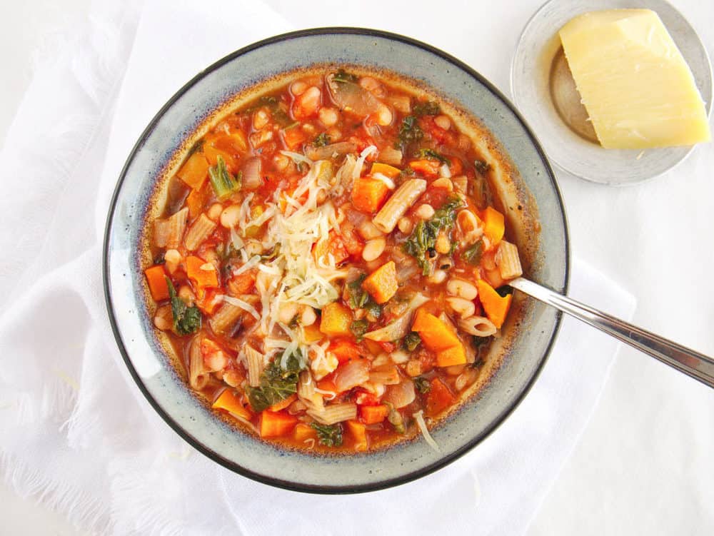 Tuscan Bean Stew with Whole Wheat Pasta, topped with parmesan cheese and served in a ceramic bowl with spoon