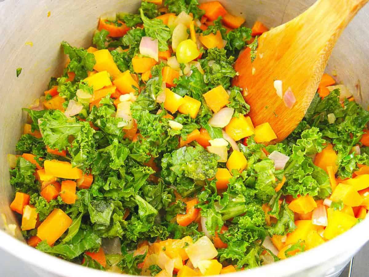 Kale added to pot with other veggies for stew in a pot on the stove.
