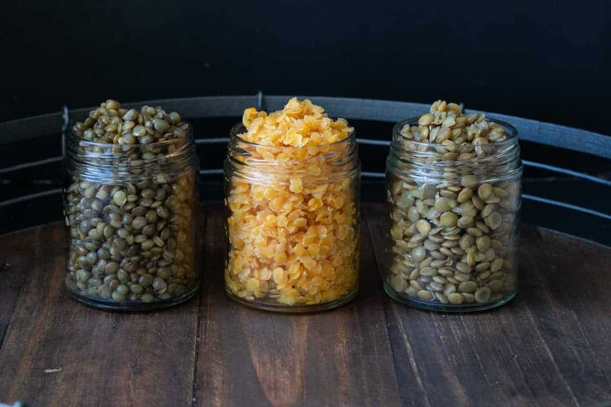 Three glass jars on a wooden tray filled with different colored cooked lentils