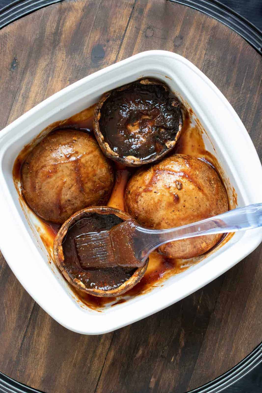 Basting brush spreading a red sauce over portobello mushrooms caps in a white dish