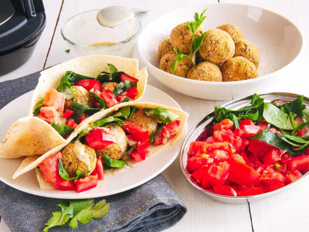 falafel wrap recipe with tomatoes, spinach and tahini sauce served on a white plate with falafel balls in the background