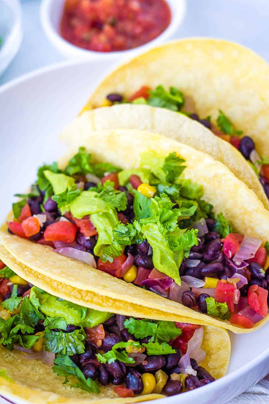 vegan tacos  with lots of taco toppings -- black beans, avocado, veggies, and cilantro, served on a white plate
