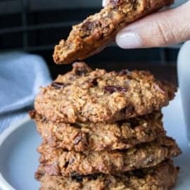 hand holding half eaten oatmeal raisin cookie with a stack of cookies on white plate