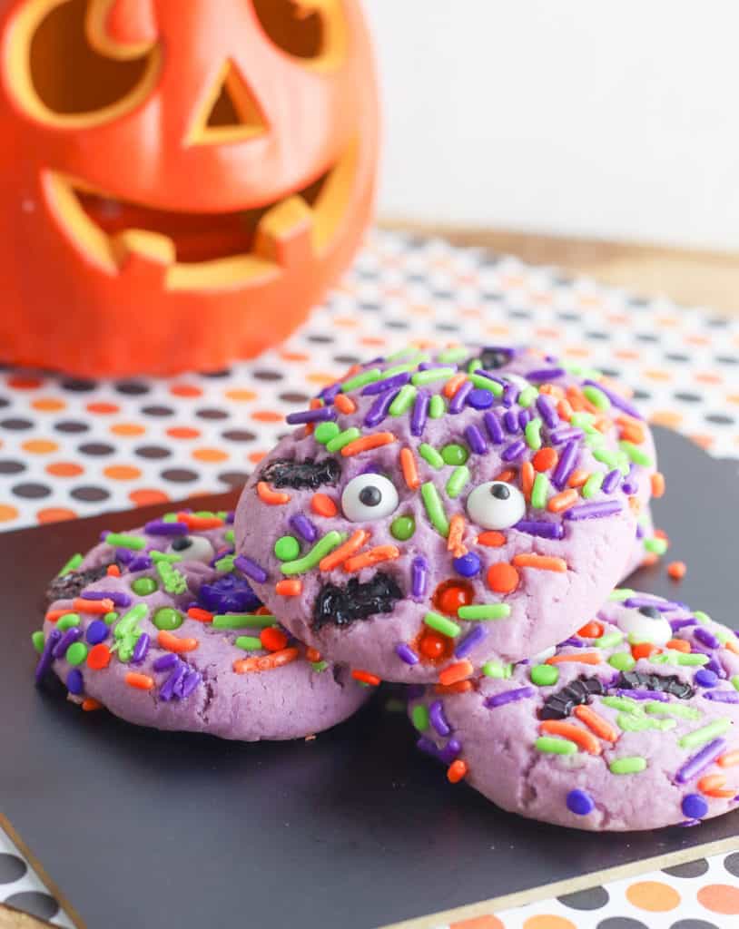 purple halloween monster cookies with googley eyes stacked on a black cutting board
