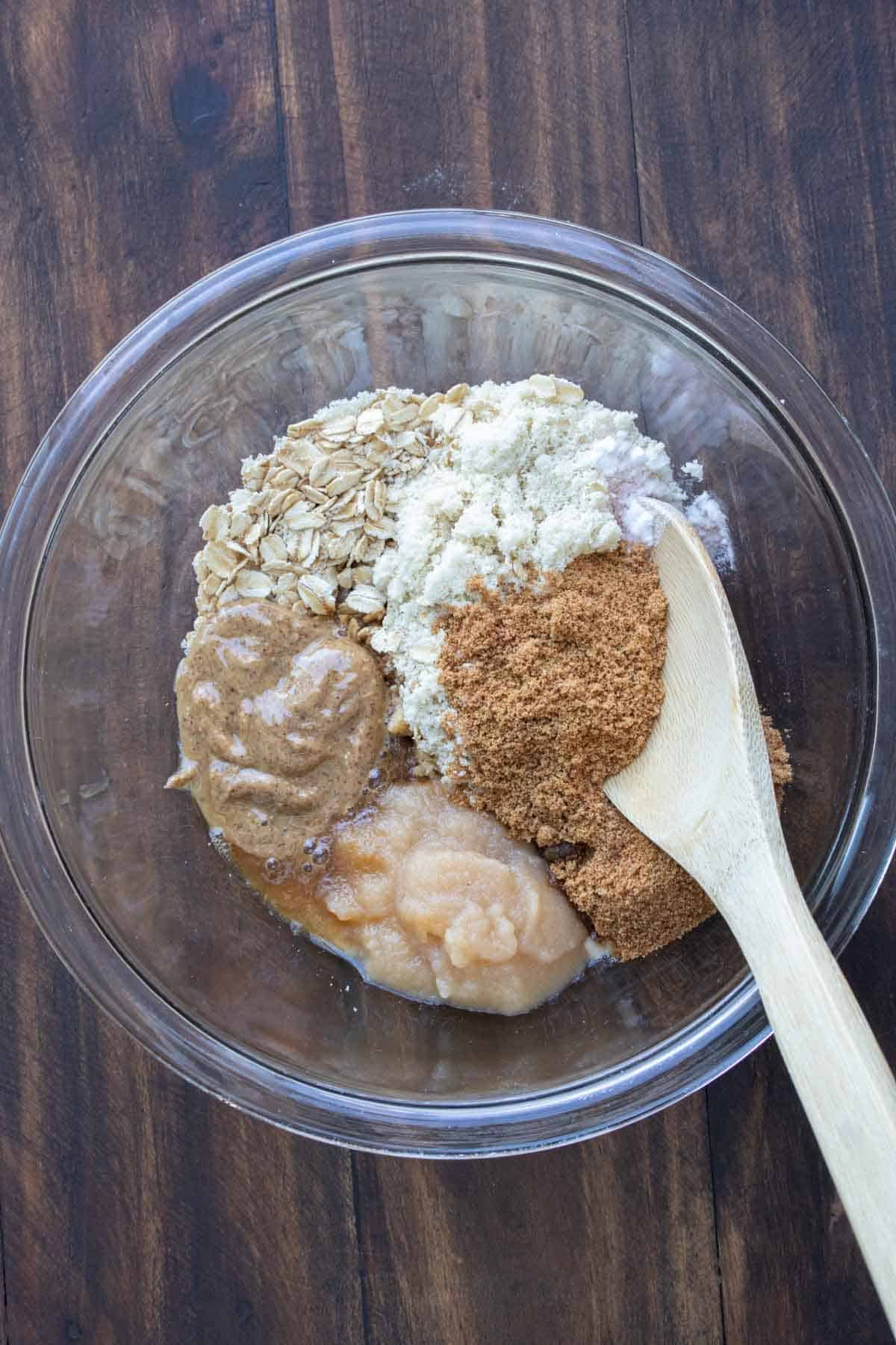 Wet & dry ingredients for oatmeal raisin cookies in a clear glass bowl with wooden spoon.