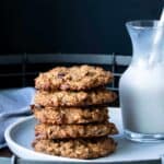 Vegan gluten free oatmeal raisin cookies stacked on a white plate next to a pitcher of milk.